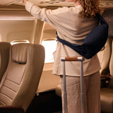 Woman retrieving luggage in an airplane, wearing a cream top, navy travel pillow, and standing next to a suitcase.